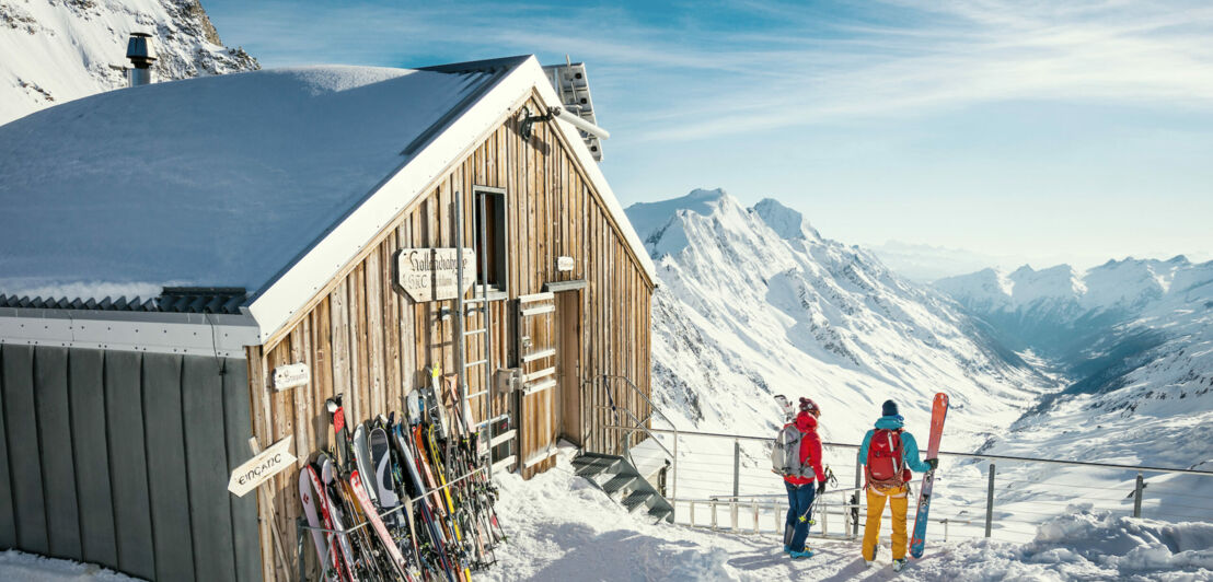 Zwei Personen stehen neben einer Holzhütte an einem Aussichtspunkt in schneebedeckter Bergkulisse