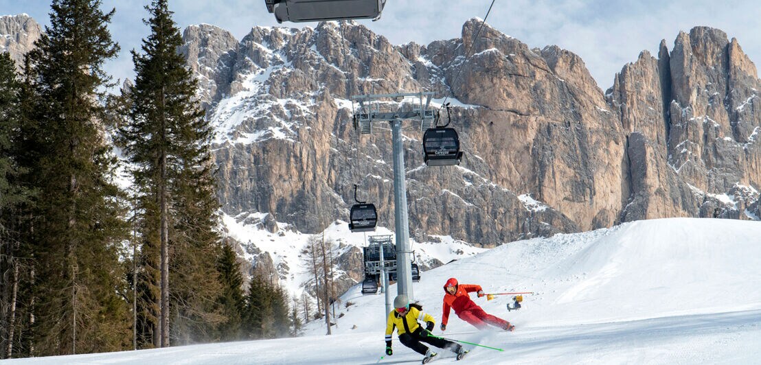 Zwei Skifahrende fahren unter einer Kabinenbahn auf einer Piste vor einem imposanten Bergmassiv