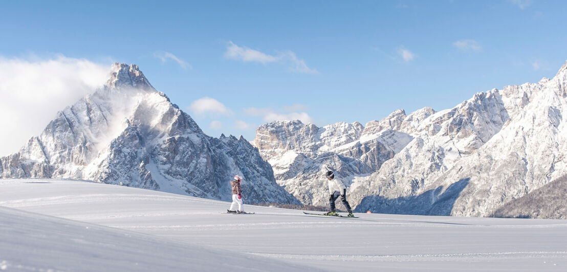 Ein Erwachsener und ein Kind auf Skiern auf einer Piste vor den schneebedeckten Gipfeln eines Bergmassivs