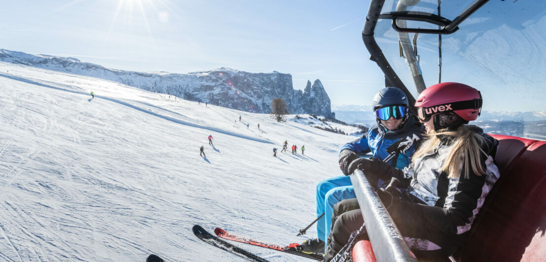 Ein Skifahrer und eine Skifahrerin sitzen in einem Sessellift über einer weitläufigen Piste vor Bergpanorama bei Sonnenschein