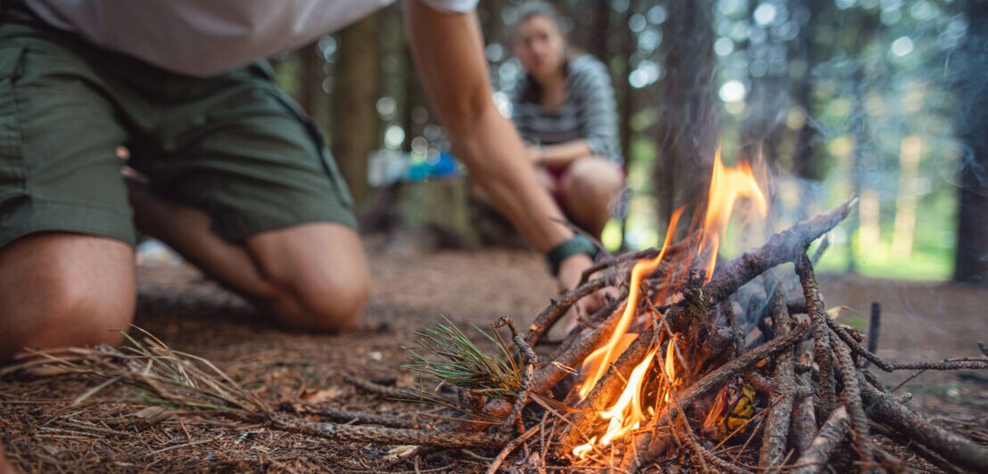 Zwei Personen hocken vor einer brennenden Feuerstelle aus Ästen in einem Wald