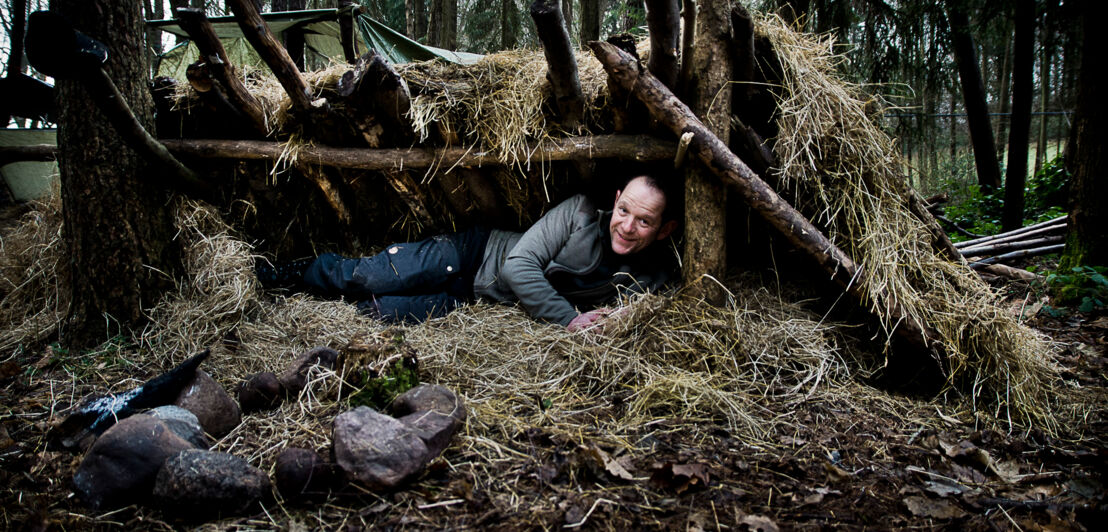Ein Mann liegt in einem Unterschlupf aus Baumstämmen und Heu in einem Wald