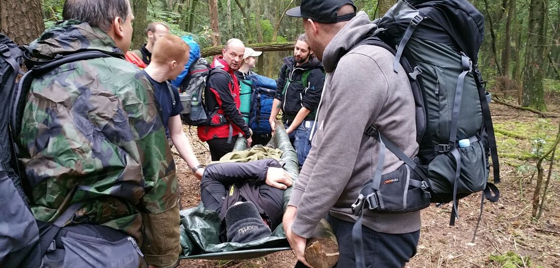 Eine Gruppe von Männern mit Rucksäcken trägt eine verletzte Person auf einer Tragbahre durch den Wald