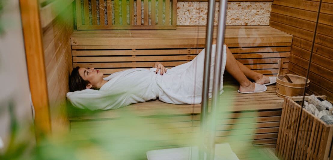 Blick durch eine Glastür auf eine junge Frau, die im Bademantel entspannt in einer Holzsauna liegt