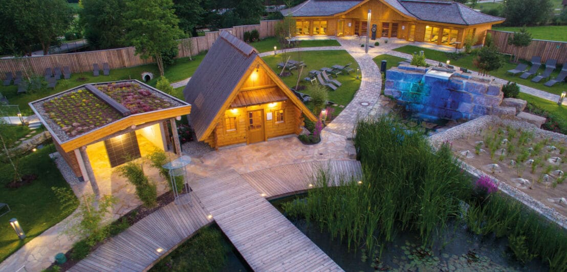 Ein idyllischer, begrünter Saunagarten mit beleuchteten Holzhütten an einem Badeteich in der Dämmerung