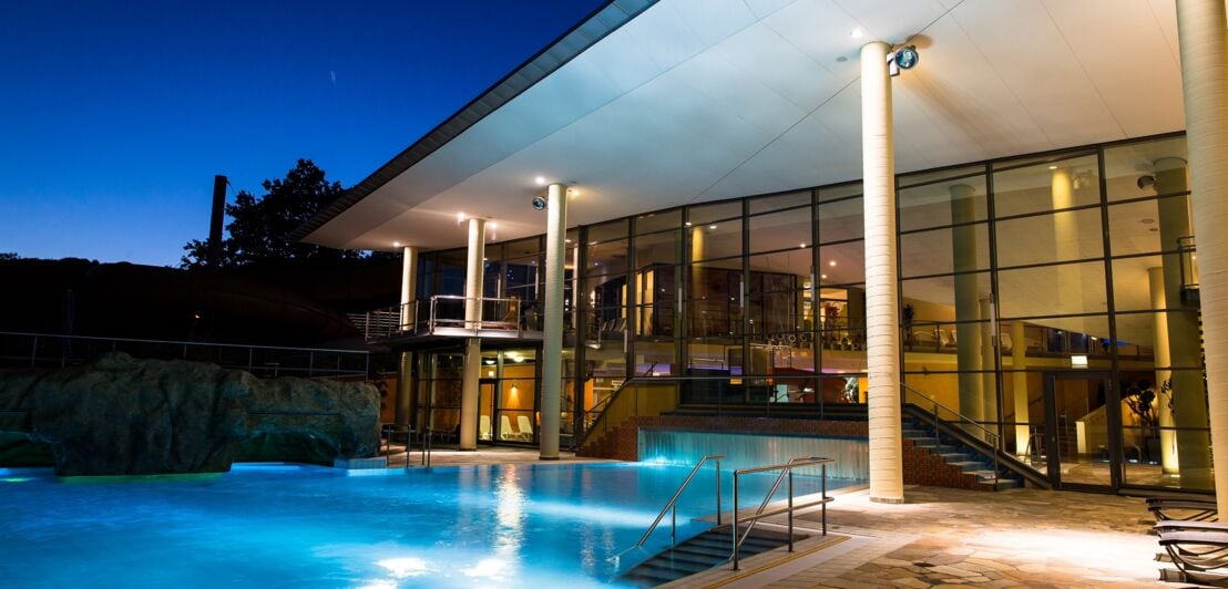 Ein beleuchtetes, geradliniges Gebäude aus dem ein Pool in den Außenbereich mit einer Terrasse führt bei Nacht
