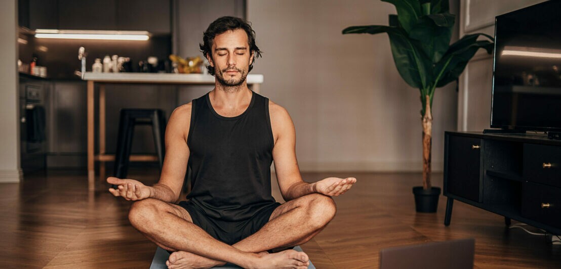 Mann sitzt zuhause auf einer Sportmatte und macht Yoga