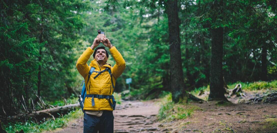 Ein Wanderer fotografiert in einem Wald mit seinem Handy in Richtung Himmel