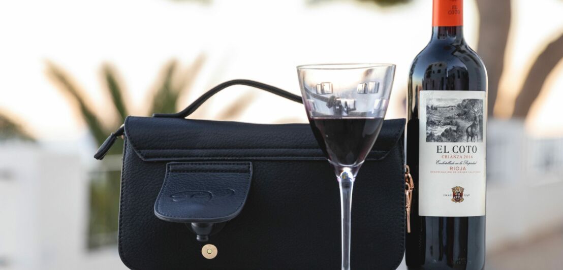 Eine Tasche mit kleiner Seitentasche inklusive Zapfhahn, daneben eine Weinflasche mit Glas