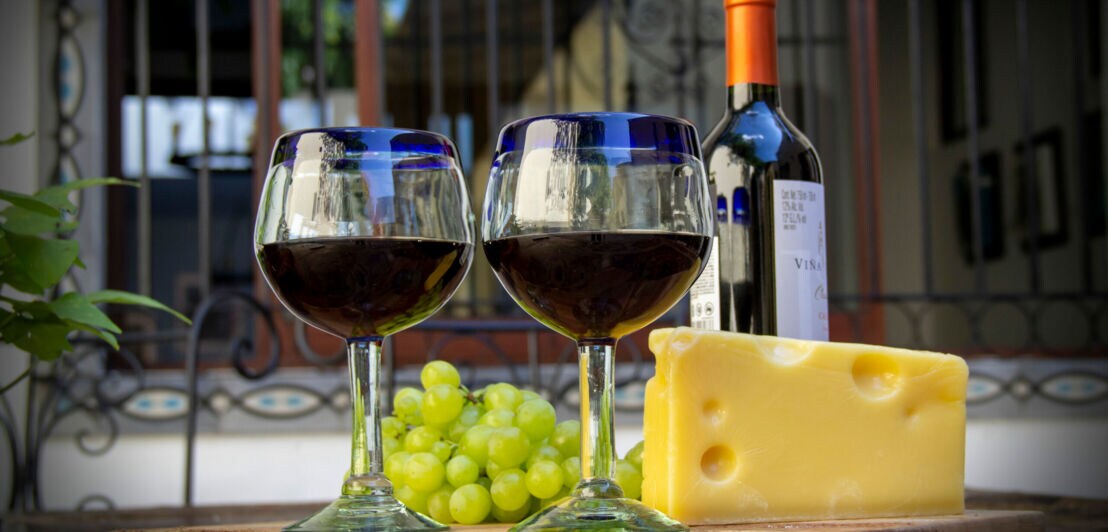 Zwei Rotweingläser sind arrangiert neben einem Stück Käse und Weintrauben