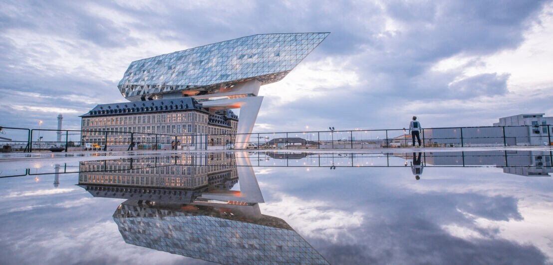 Ein futuristischer Neubau auf einem alten Gebäude, davor Wasser