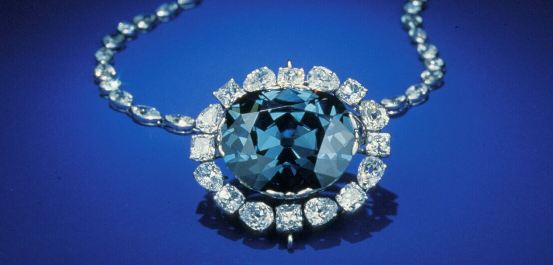 Ein großer, blauer Diamant, eingefasst in kleinere Diamanten an einer Kette auf blauem Untergrund
