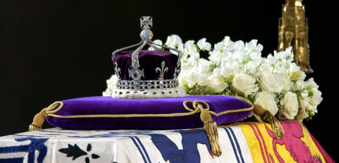 Eine diamantenbesetzte Krone auf einem Kissen, dahinter ein Blumengesteck