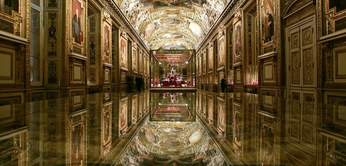 Innenaufnahme eines prunkvollen Saals im Louvre mit Ölgemälden und Fresko, im Zentrum eine Vitrine mit Schmuck