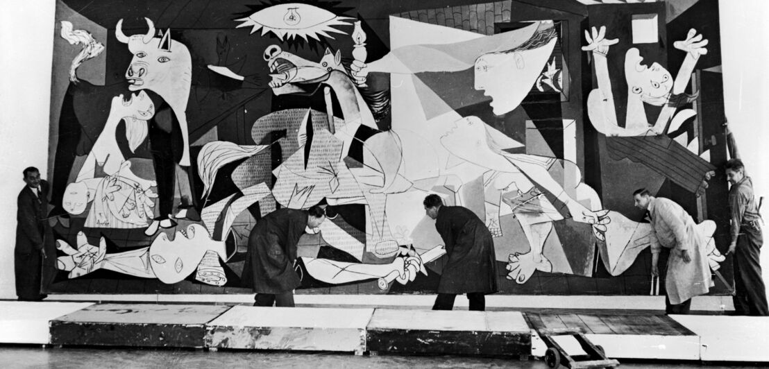 Historische Schwarzweißaufnahme von Picassos Monumentalgemäldes Guernica, das von vier Männern von der Wand genommen wird