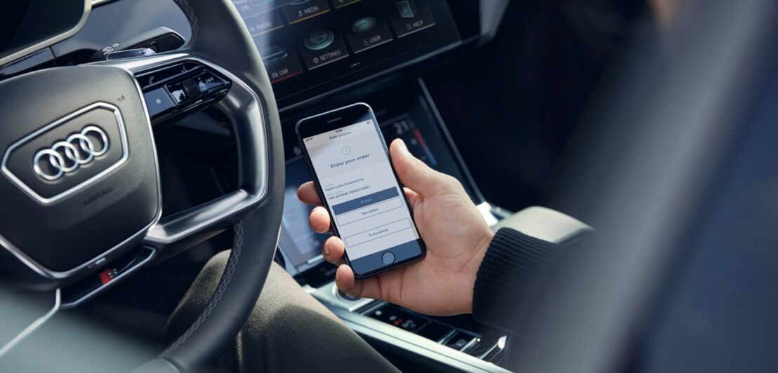 Fahrer hält im Cockpit eines Audis ein Smartphone in der Hand, um eine Funktion für das Auto zu buchen
