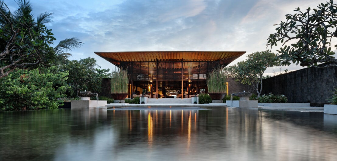 Ein halboffener, luxuriöser Pavillon in puristischem Design, umgeben von tropischen Bäumen und einem Wasserbecken