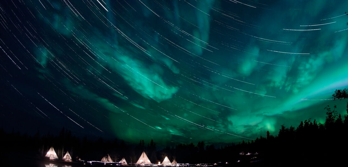 Grüne Polarlichter am Nachthimmel, unter ihnen ein beleuchtetes Tipi-Dorf