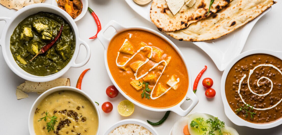Aufsicht einer Auswahl an vegetarischen, indischen Gerichten in weißen Schalen auf weißem Untergrund