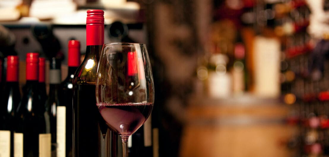 Ein Glas mit Rotwein steht auf einem Holztisch, im Hintergrund Weinfalschen