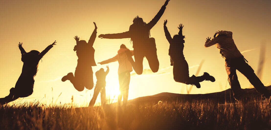 Gegenlichtsituation: Eine Gruppe junger Menschen springt mit erhobenen Armen in die Luft, auf einem Grashügel vor der untergehenden Sonne