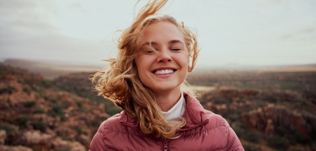 Porträt einer jungen, blonden Frau vor karger Felslandschaft, sie lächelt und hat die Augen geschlossen