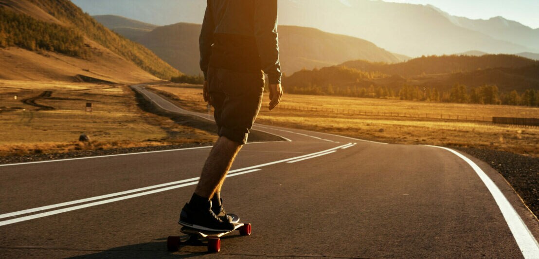 Karge Landschaft vor einer Bergkette, ein Mann fährt auf einem Longboard eine lange, leere Straße entlang.