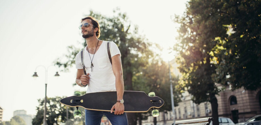 Mann mit Sonnenbrille hält ein Longboard, dahinter Stadtkulisse