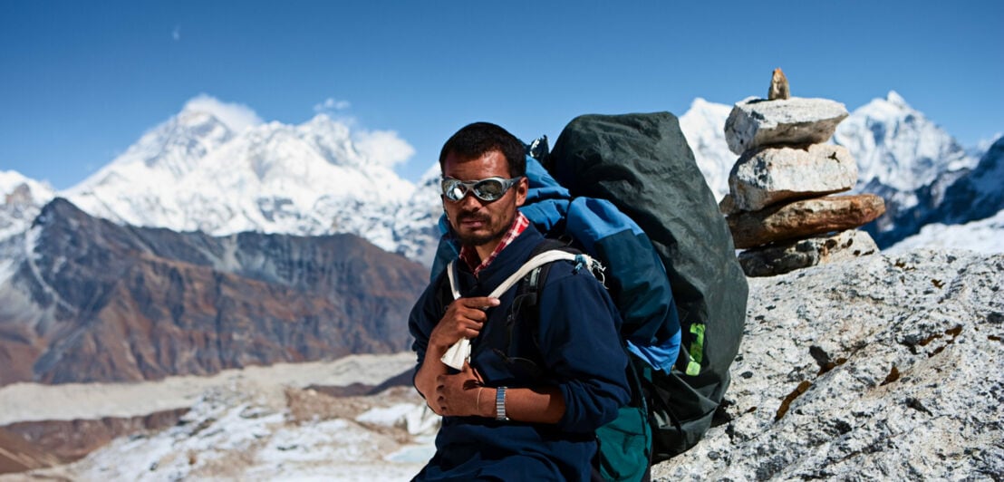 Ein Sherpa mit Rucksack lehnt an einem Felsen, im Hintergrund Berge