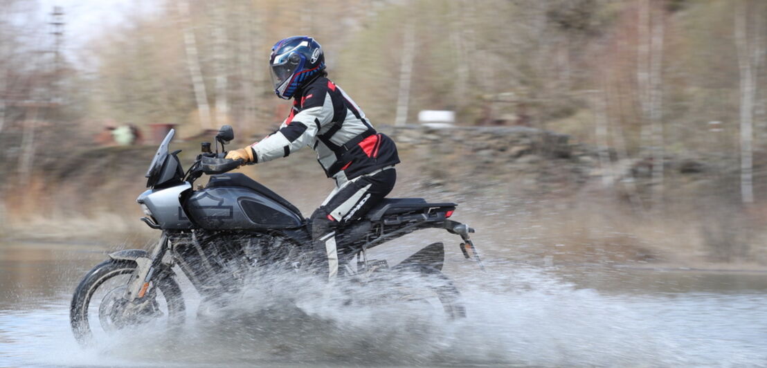 Ein Motorradfahrer in Endurobekleidung fährt auf einer grauen Harley-Davidson durch ein Flussbett und lässt Wasser aufspritzen.