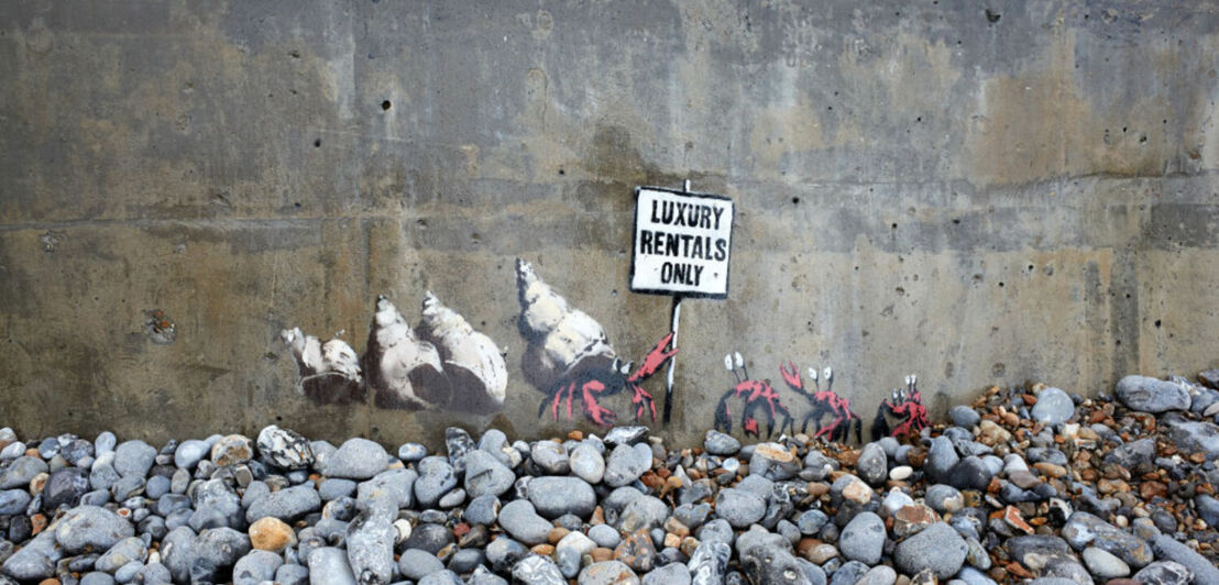 Ein Werk des Künstlers Banksy, das einen Krebs zeigt, der mehrere Schneckenmuscheln vor anderen Krebsen schützt und ein Schild mit der Aufschrift „Luxury Rentals Only“ hochhält.