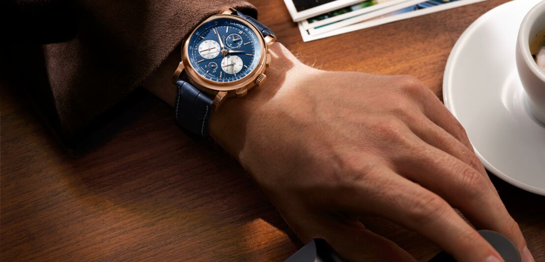 Close-up einer in Blau gehaltenen Uhr von A. Lange und Söhne an einem Männerarm, der auf einem Holztisch mit Fotokamera und Espressotasse liegt