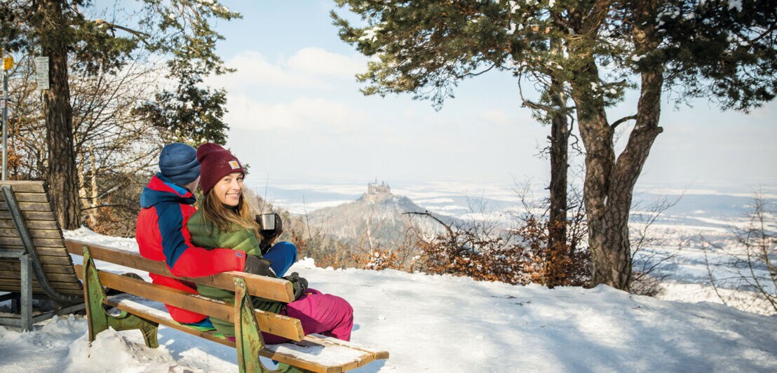 Ein Paar sitzt auf einer verschneiten Bank und genießt den Blick in die umliegende weiße Winterlandschaft mit einem Thermobecher in der Hand