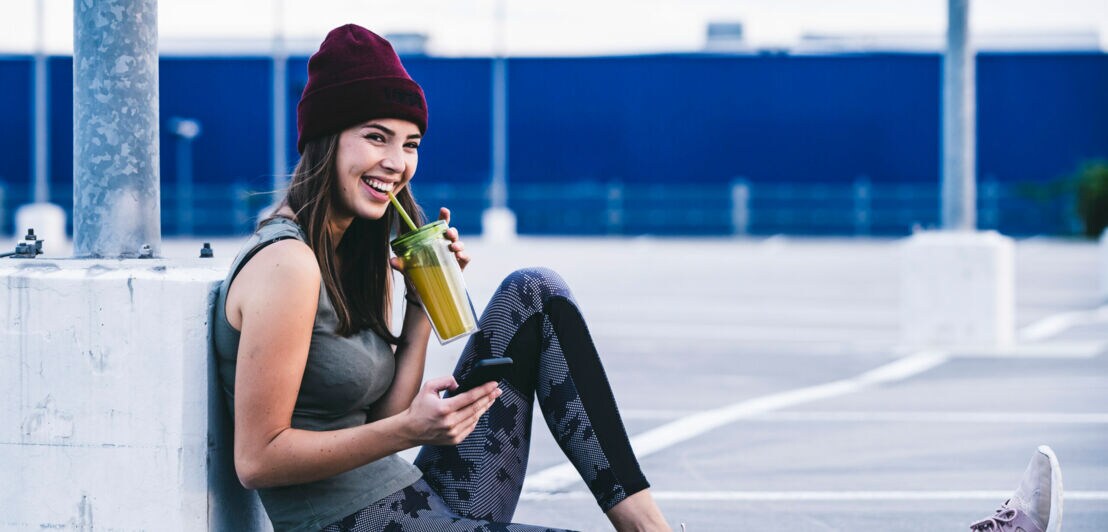 Eine junge Frau in Sportkleidung trinkt einen Smoothie, neben ihr liegt eine Hantel.