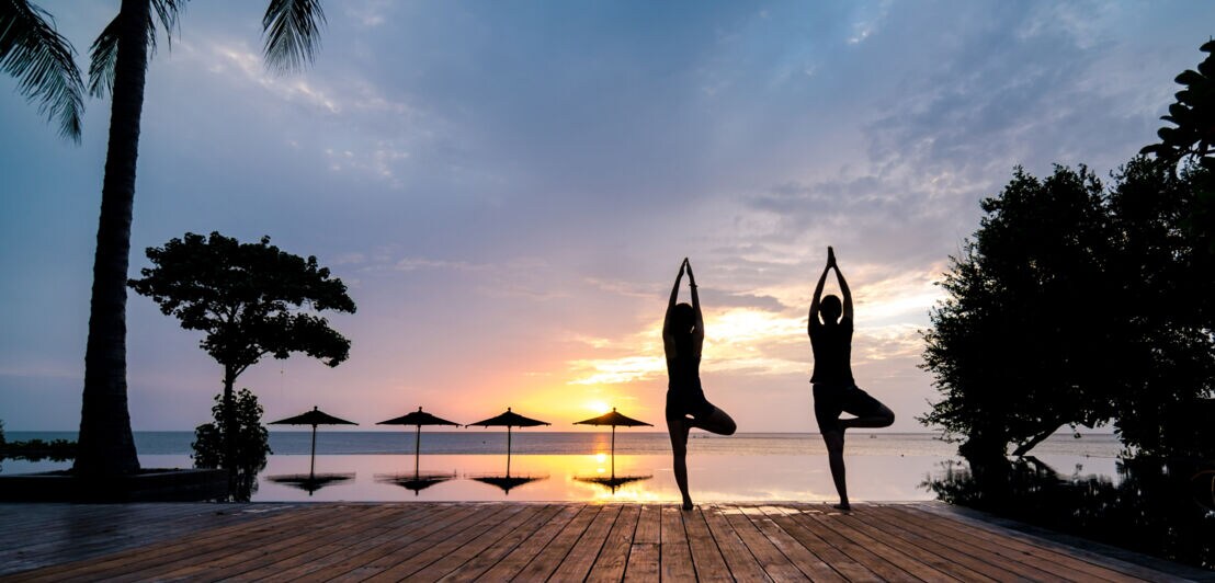 Silhouetten von zwei Personen, die im Sonnenuntergang an einem Hotelpool mit Blick aufs Meer Yogaübungen machen