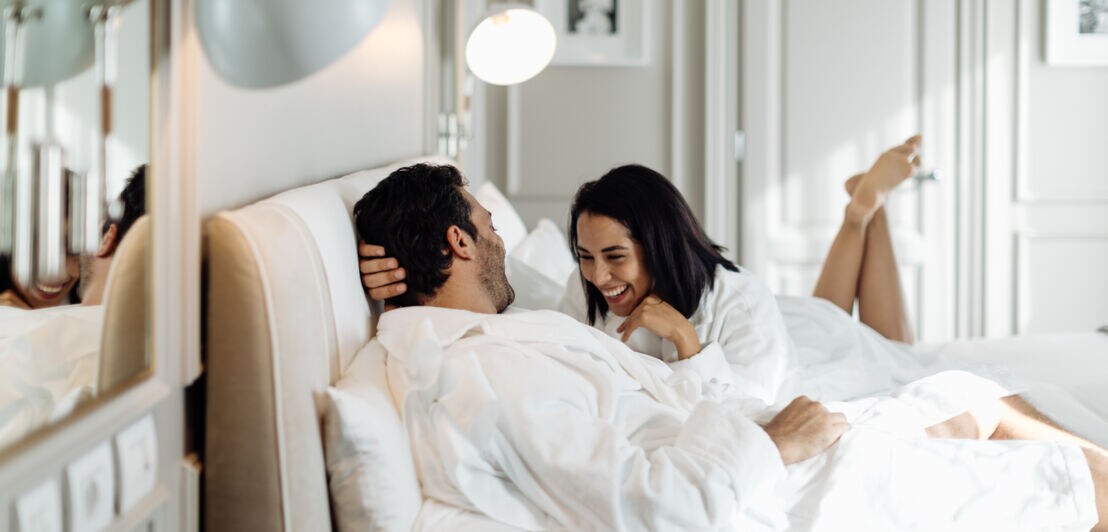 Ein Paar in Bademänteln liegt lachend auf dem Bett in einem hellen, modernen Hotelzimmer