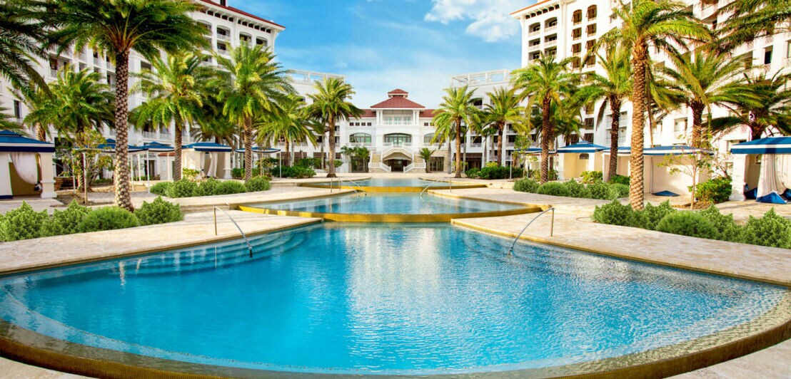 Ein großer Pool mit vielen Palmen zwischen zwei Hotelkomplexen.