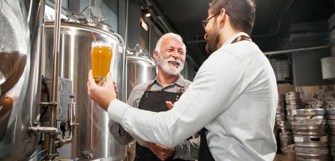 Zwei lächelnde Männer in einer Brauerei, von denen einer ein Glas Bier hochhält