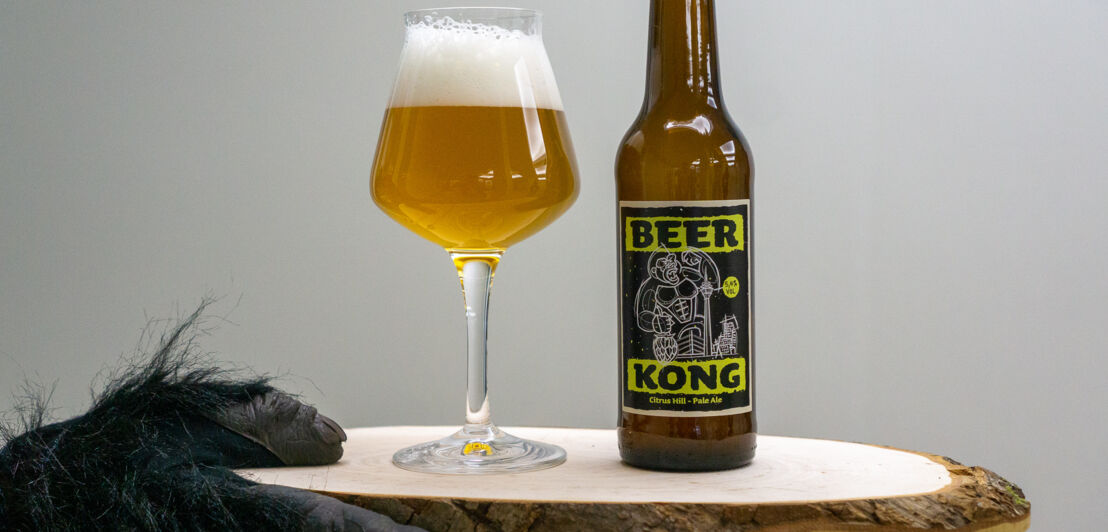 Ein Glas Bier und eine Flasche auf einer Holzplatte, die von einer Hand in einem Affenkostüm gehalten wird