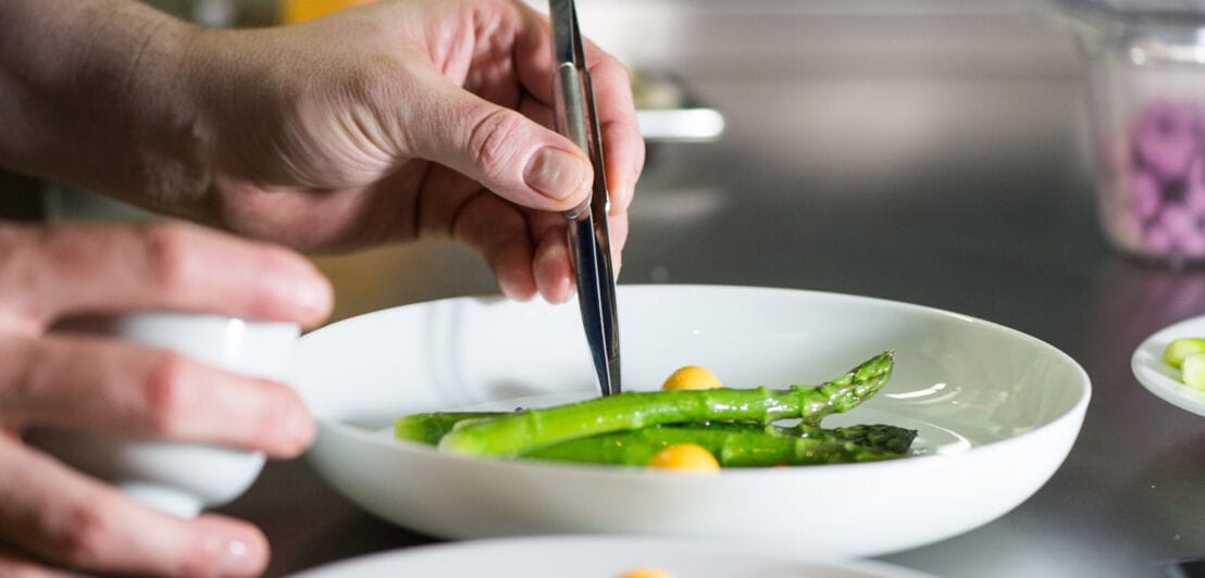 Nahaufnahme einer Hand, die mit einer Pinzette grünen Spargel und Mangostücke auf einem weißen Teller in einer Küche anrichtet