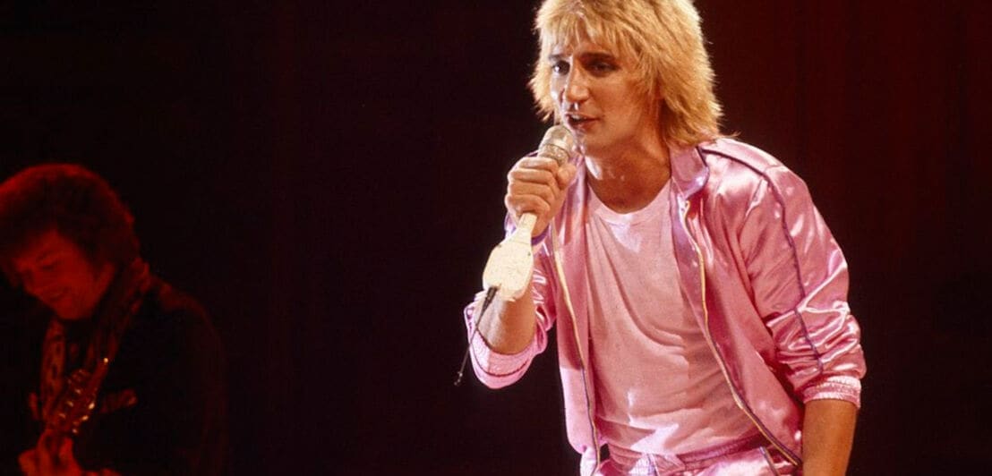 Der Sänger Rod Stewart trägt einen pink-glänzenden Jogginganzug und einen wuscheligen Haarschnitt