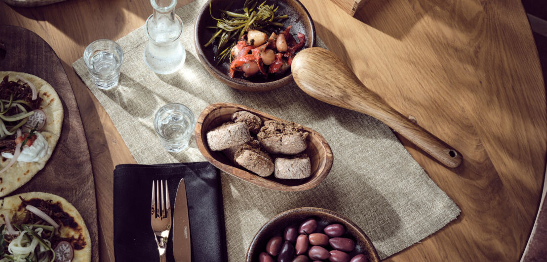 Ein gedeckter Holztisch mit Oliven, Pizzen, Brot, Gläsern, kleinen Schalen und Besteck