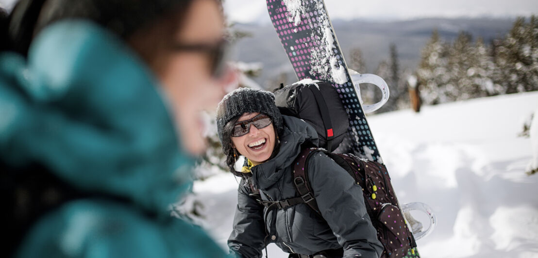 Eine lachende Frau in Winterkleidung mit einem Rucksack und einem Snowboard auf dem Rücken in einer Schneelandschaft
