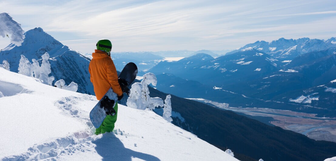 Ein Snowboarder steht mit seinem Board unterm Arm an einem Hang im Tiefschnee und blickt auf eine Berglandschaft