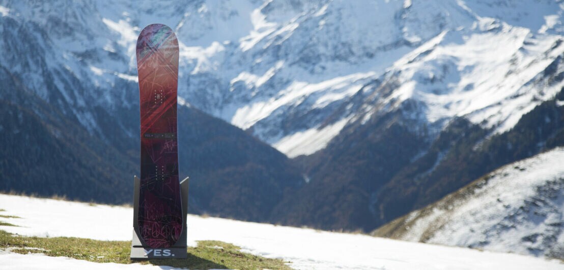 Ein Snowboard der Marke YES steht senkrecht vor einem Bergpanorma