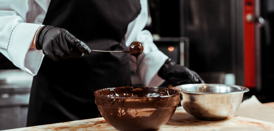 Ein Mensch mit Kochschürze hält einen schokoladigen Löffel über einer schokoladigen Schüssel