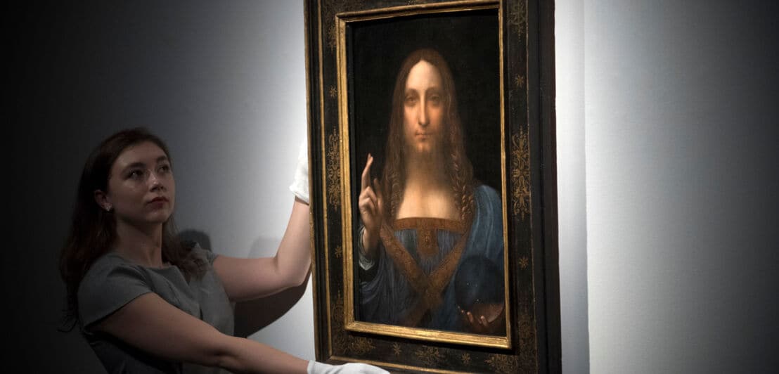 Eine Frau mit weißen Handschuhen präsentiert Leonardo da Vincis Gemälde Salvator Mundi vor einer weißen Wand