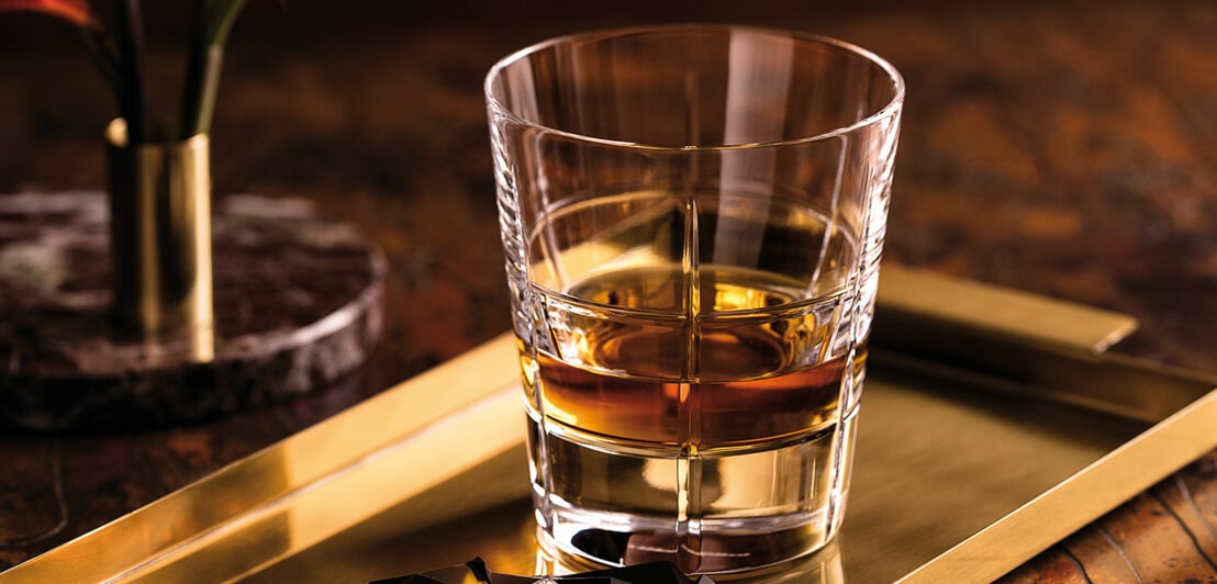 Ein leicht gefülltes Whiskyglas von Villeroy & Boch auf einem goldenen Tablett mit Bruchstücken dunkler Schokolade, dahinter steht eine kleine Vase mit zwei Montbretien