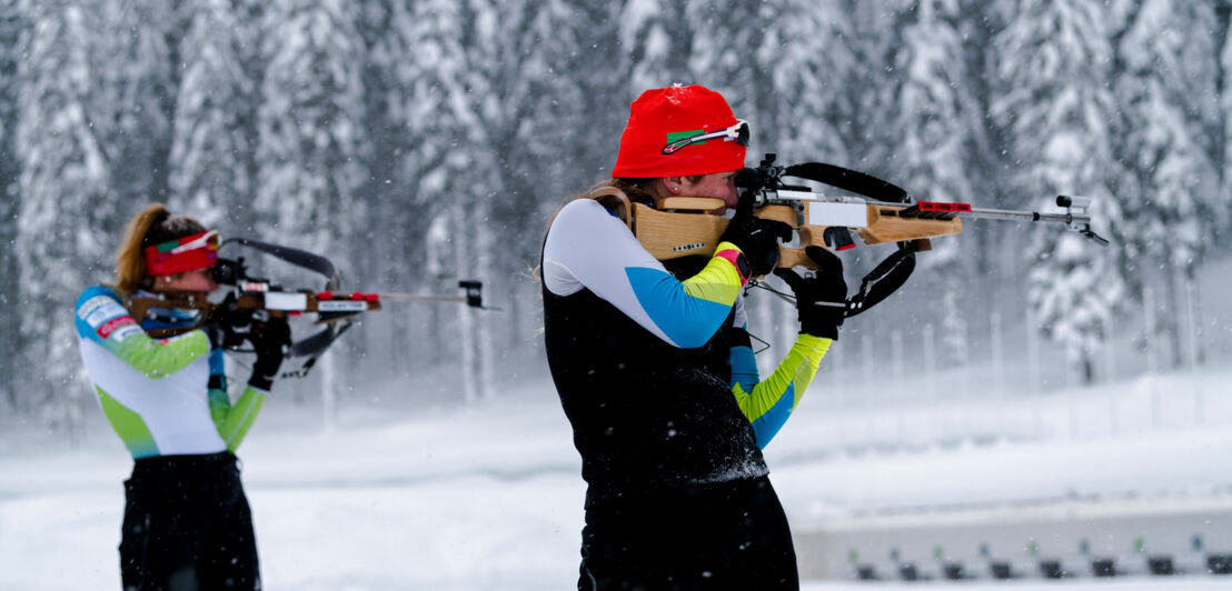 Zwei Athletinnen in sportlicher Kleidung üben Zielschießen in verschneiter Umgebung.