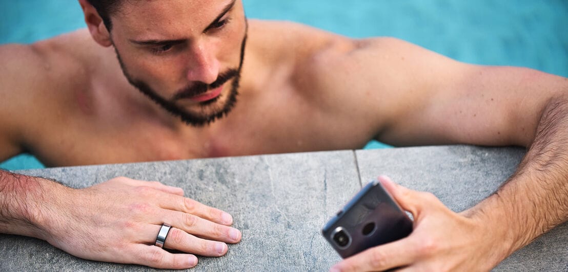 Ein Mann im Pool mit Smart-Ring am Finger schaut auf sein Handy